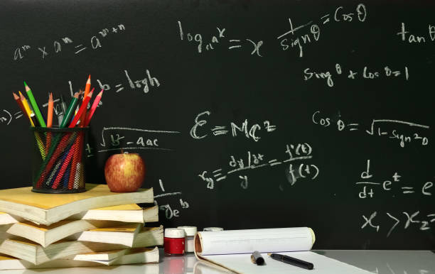 Books are stacked on table with mathematical equations on blackboard